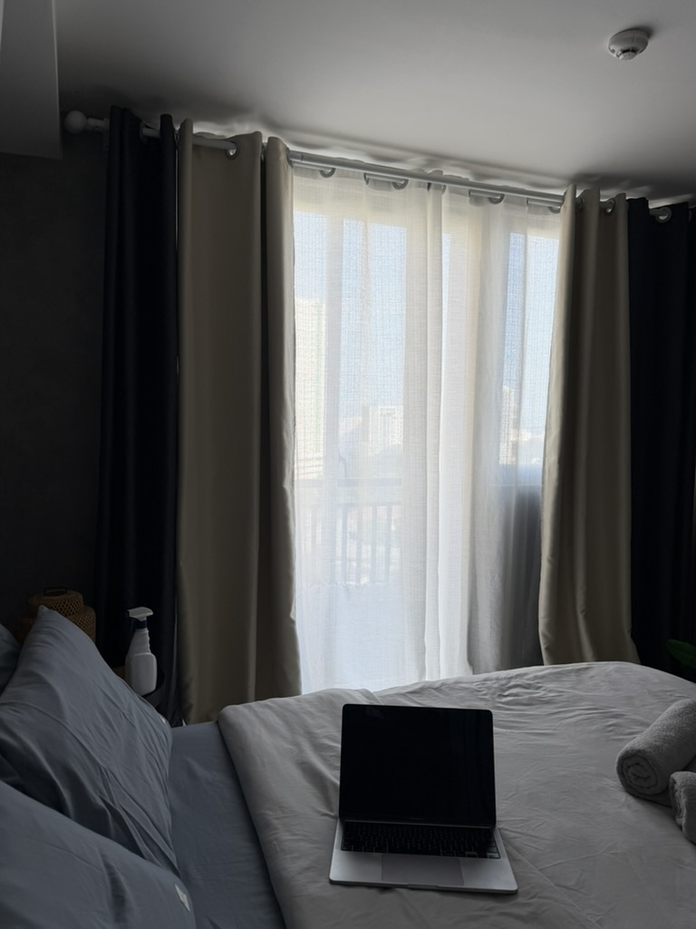 Bedroom workspace setup at Cityscape Grand Tower with laptop on the bed and sheer city-view curtains